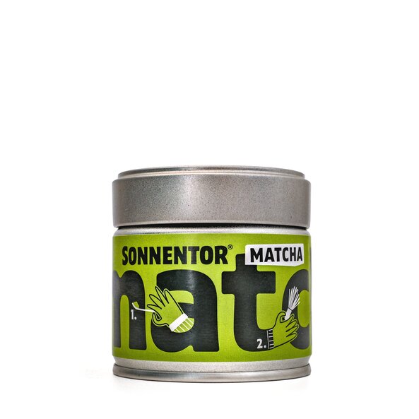 auf der kleinen Blechdose ist ein grünes Etikett mit der schwarzen Aufschrift "matcha". In den Buchstaben sind die Zubereitungsschritte mit Symbolen dargstellt. 