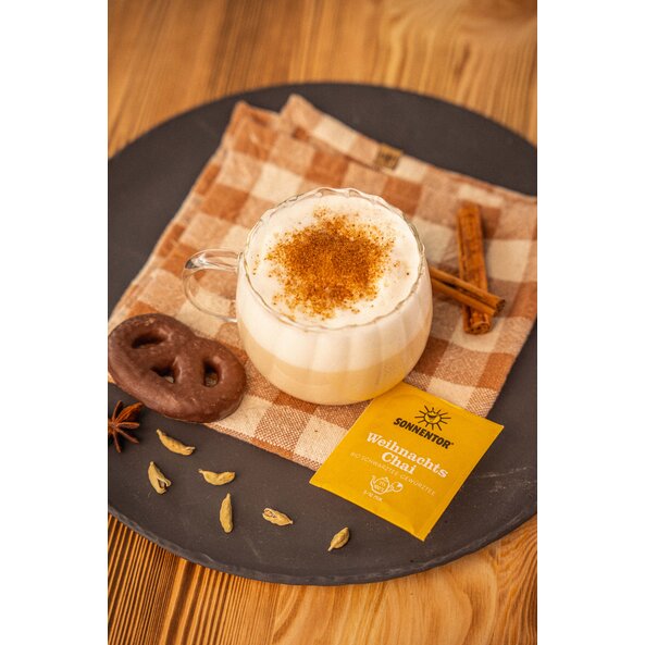 SONNENTOR Weihnachts Chai Bio neben Glas mit cremigem Chai, Zimtstangen, weiteren Gewürzen und Lebkuchen