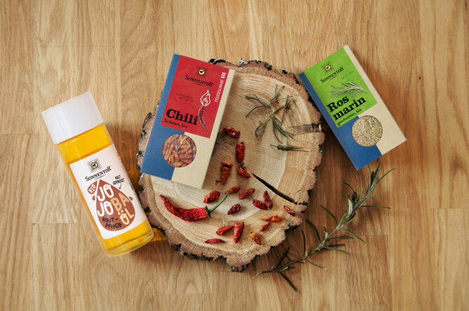 Jojoba-Rosmarin-Chili | © SONNENTOR Auf dem Foto ist eine Holzscheibe zu sehen. Darauf sieht man Chilischoten und Rosmarin. Daneben liegt eine Flache Jojobaöl. | © SONNENTOR