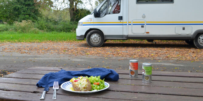 Camper | © SONNENTOR Auf dem Foto sieht man einen Tisch mit einem Teller mit Ofenkartoffel darauf. Hinter dem Tisch sieht man einen Camper. | © SONNENTOR